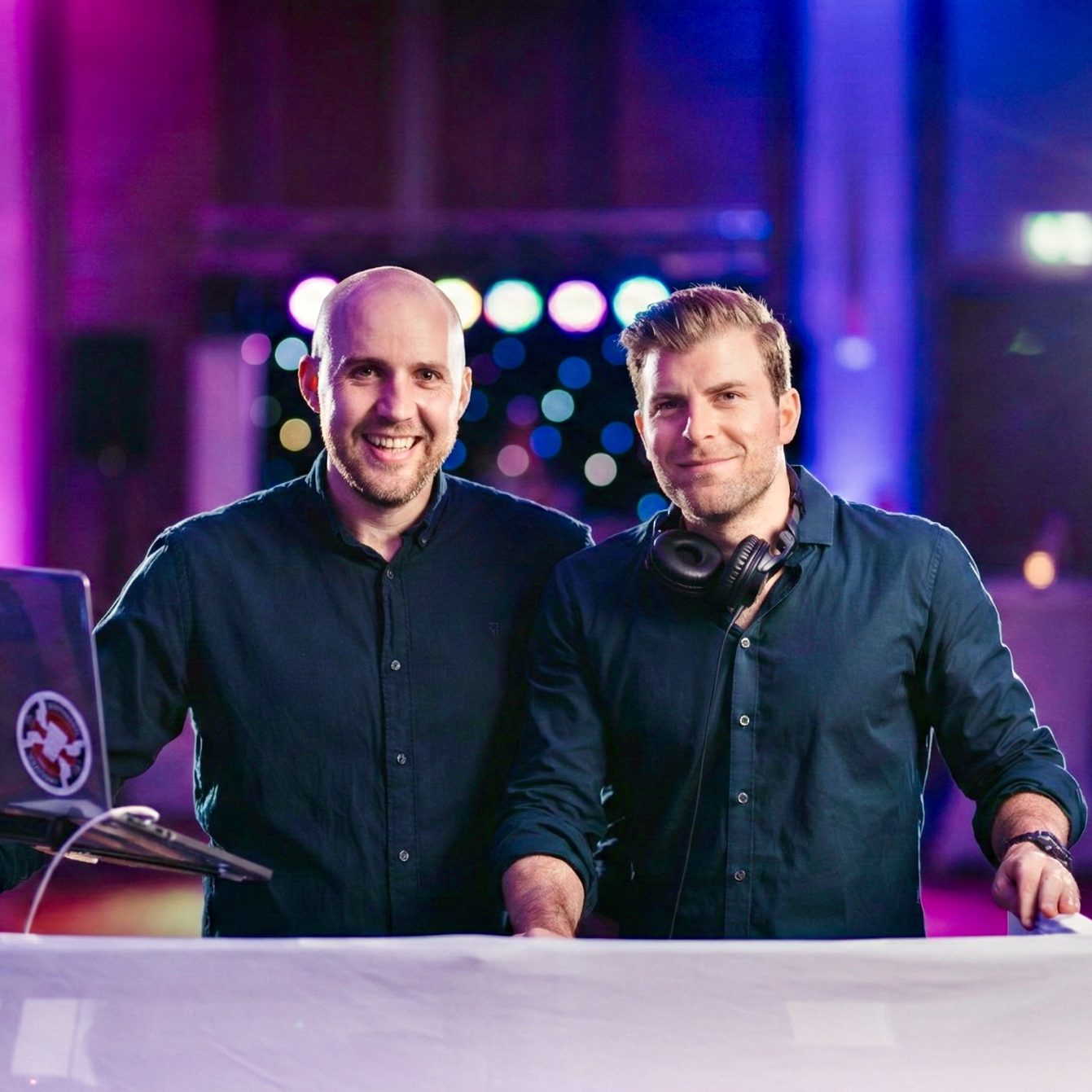 Zwei DJs stehen am Mischpult, lächeln und präsentieren ihre Ausrüstung in lebendiger Atmosphäre.